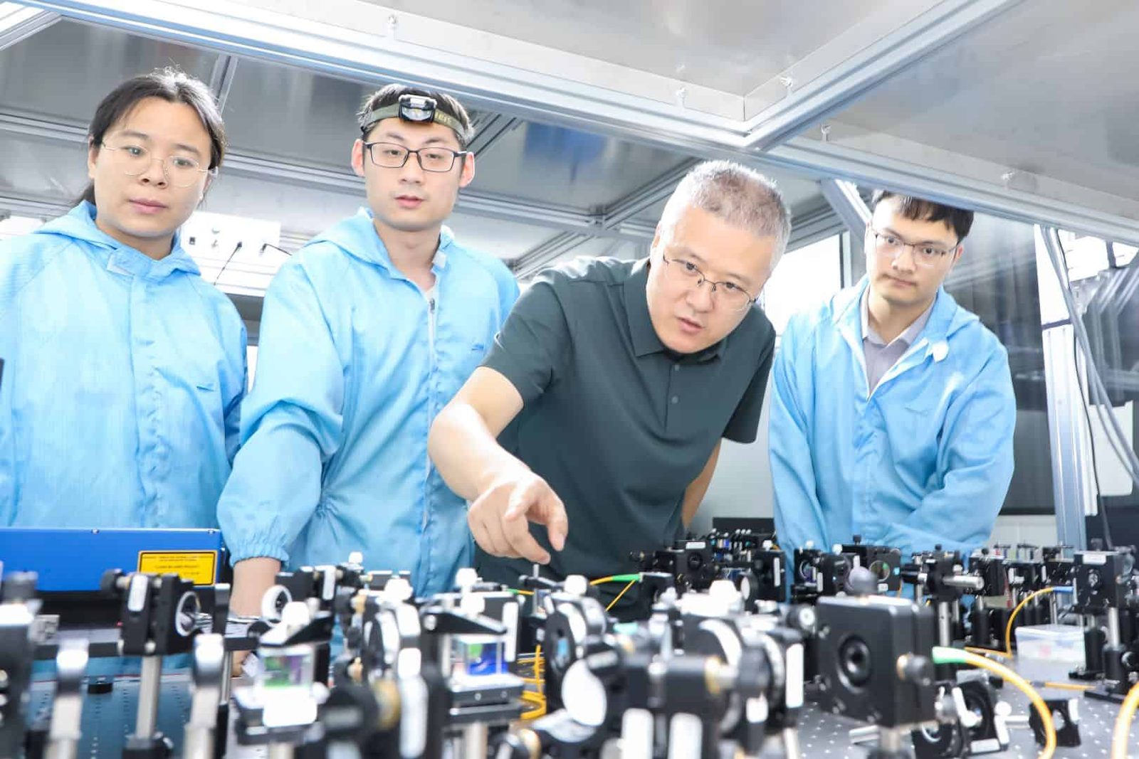 Photo of four people (Qi Zhang, Xu-Cai Zhuang, Zhong-Xiao Man and Qing-Feng Xue) standing next to an optical bench. Man is wearing a button-up polo shirt and pointing an an optical element on the table. The other three are wearing blue clean-room suits. Zhuang wears a head lamp and appears to be adjusting something on the table